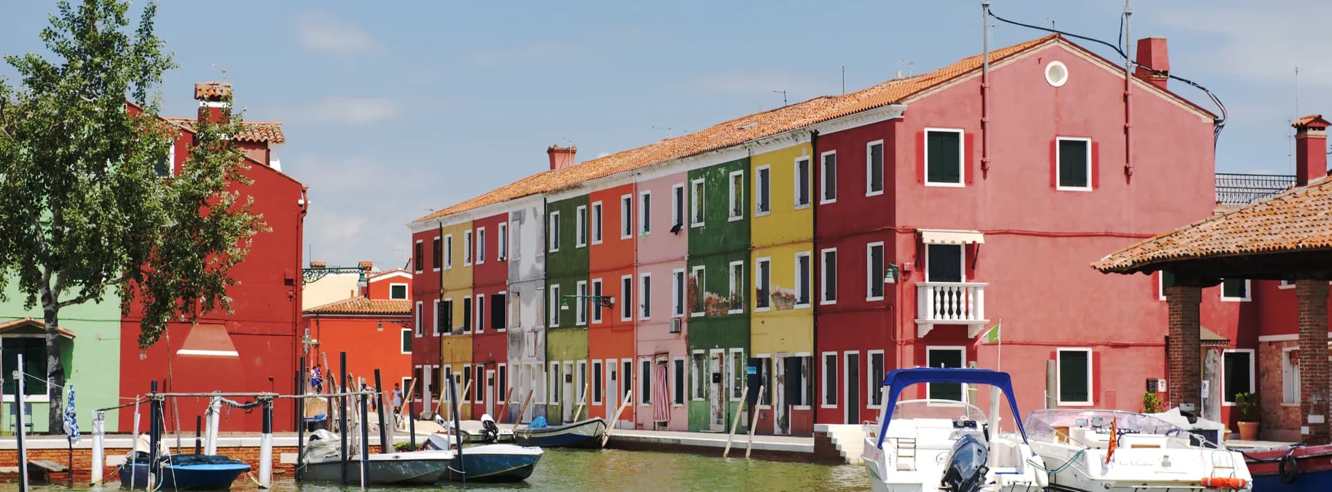 View approaching a canal in Burano, Italy with anchored boats and a row colorful buildings